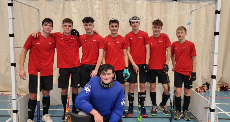 Ashby school hockey team posing in front of a goal net, wearing grey uniforms with some players kneeling and others standing, with a coach in a blue jacket
